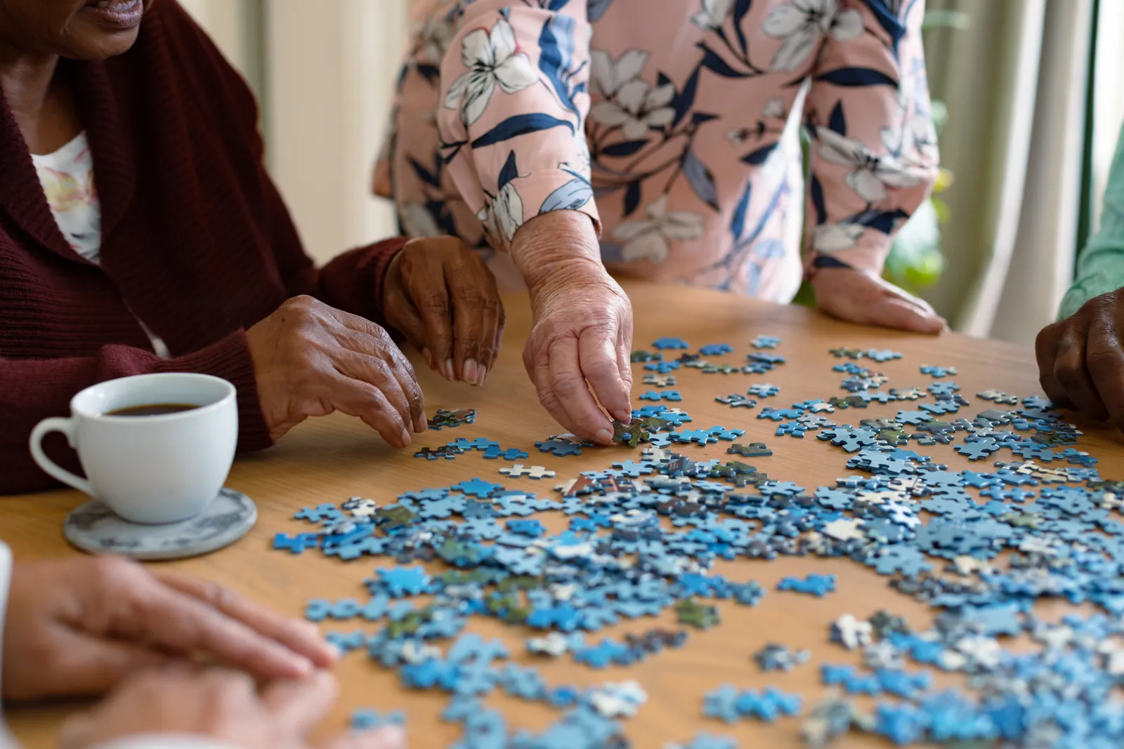 Seniors' hands doing a blue jigsaw puzzle on a table beside a cup of tea