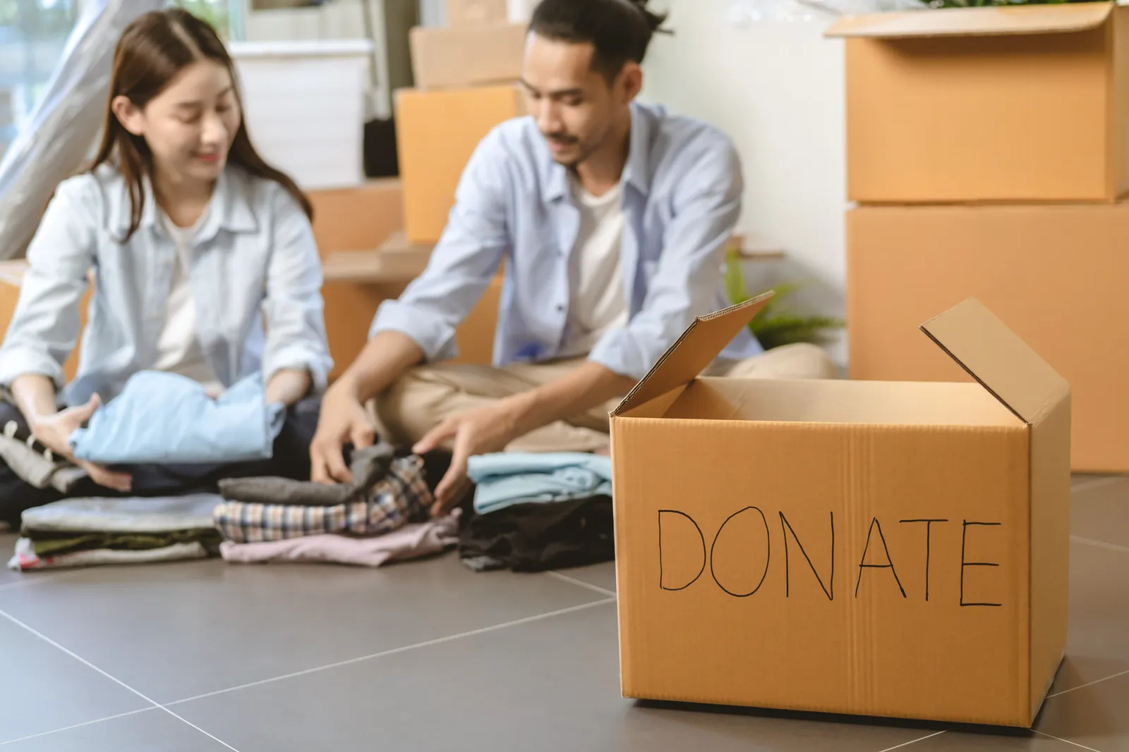 Winter Wardrobe Detox. A women and a man sitting on the ground sorting through clothes. Cardboard boxes around then for their winter wardrobe detox, sorting into piles to Keep, Donate, Recycle